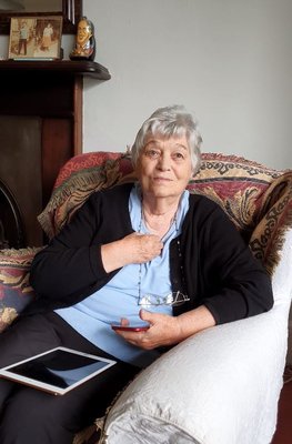 Imelda is sitting in an arm chair, wearing a blue shirt and black cardigan, an iPad rests of her lap.