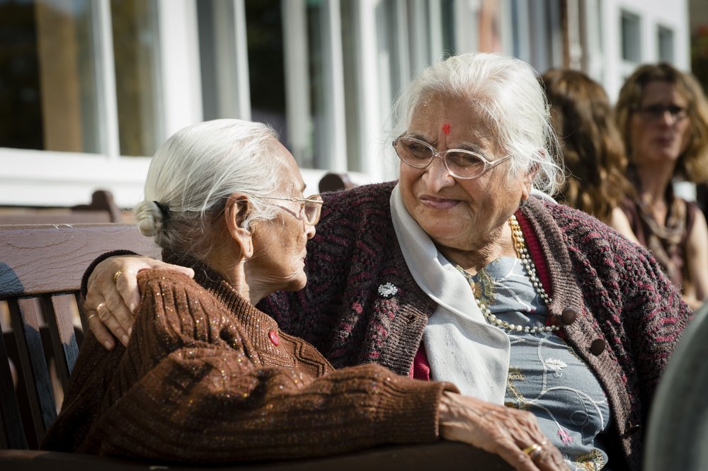 A picture of two ladies sat together at a bench, smiling and looking at each other.