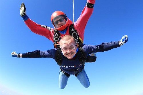 Two people skydiving, they are both smiling and have their arms out to their side. One person is wearing a t-shirt which says 'Vista'.
