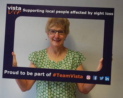 Jean Voller standing and smiling with a Vista branded selfie frame which says 'Supporting local people affected by sight loss. Proud to be part of #TeamVista'. This copy is followed by the logo for Facebook, Twitter and Instagram at the bottom right-hand side.