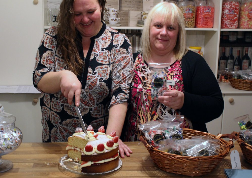A picture of two care staff stood at the sweet shop counter, cutting a slice of cake.