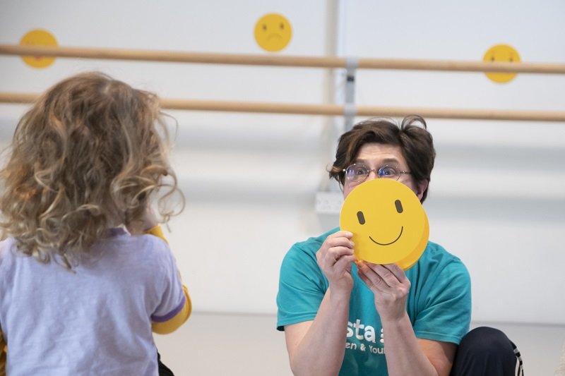 Image is of a child looking at Oksana Tyminska Vista’s Early Years Project Manager, holding a circle of yellow paper with a smiley face drawn on it.