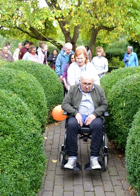 An elder man in a wheelchair being pushed by a smiling carer through gardens