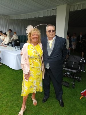 Image is of Julie Rudd in a yellow dress standing smiling next to John under at tent at the Buckingham Palace garden party.
