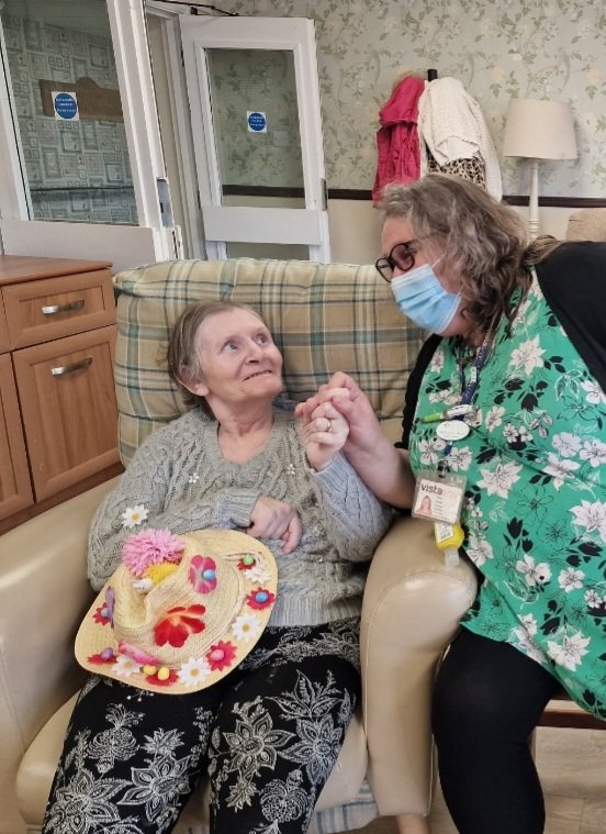 A member of staff at Vista's New Wycliffe Home sitting with a resident and they are smiling. The resident is holding a decorated Easter bonnet.