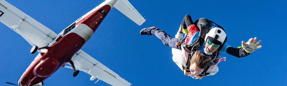 Image Description: A man smiles and reaches towards the camera as he jumps from a plane on a skydive, with another person attached to him.