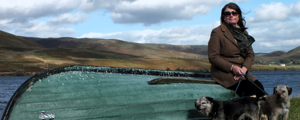 [Image Description: Chrissy wears a coat and sunglasses as she sits on the edge of an overturned boat beside a lake, holding the leads of two dogs. Rolling hills and cloudy skies stretch across the background, creating a calm outdoor scene.]