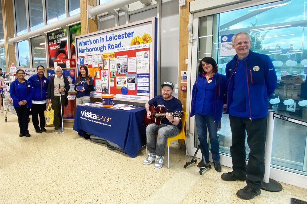 The Blind Guitar Guy, Craig, Jemima and TESCO Colleagues gather around a Vista information stall hosted in a TESCO store. Everyone is smiling and happy.