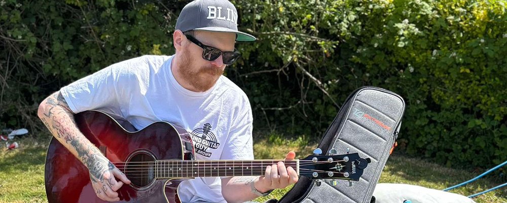 [Image Description: Craig, known as the Blind Guitar Guy, sits on a hay bale playing an acoustic guitar outdoors on a sunny day. He wears a cap with the word “BLIND” on it, dark sunglasses, and a branded t-shirt. Next to him is a grey guitar case with a custom “Blind Guitar Guy” patch.]