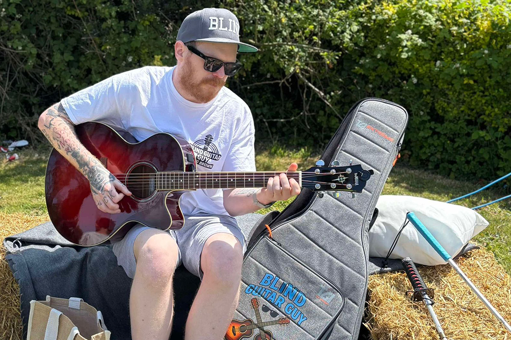 [Image Description: Craig, known as the Blind Guitar Guy, sits on a hay bale playing an acoustic guitar outdoors on a sunny day. He wears a cap with the word “BLIND” on it, dark sunglasses, and a branded t-shirt. Next to him is a grey guitar case with a custom “Blind Guitar Guy” patch.]