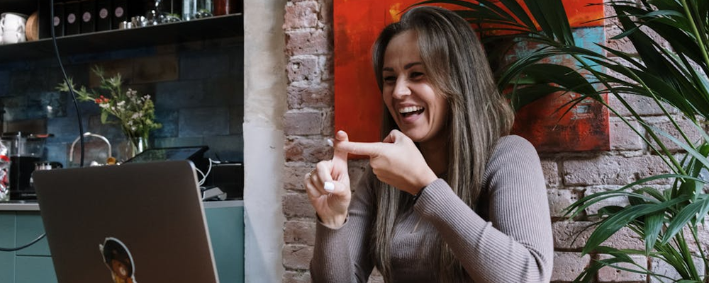 [Image Description: Woman in Gray Sweater Sitting by the Table Doing Hand Sign While Having Video Chat]
