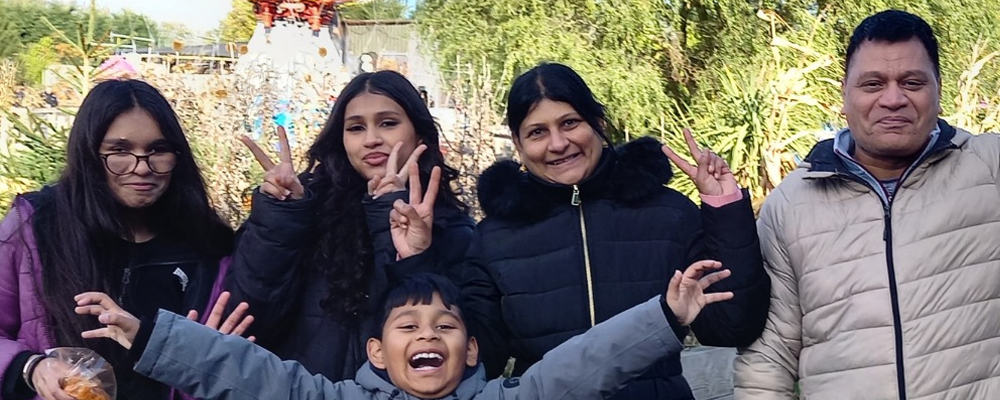 [Image Description: A family of five poses happily at an amusement park, with a thrill ride visible in the background. The young boy in front is smiling widely.]