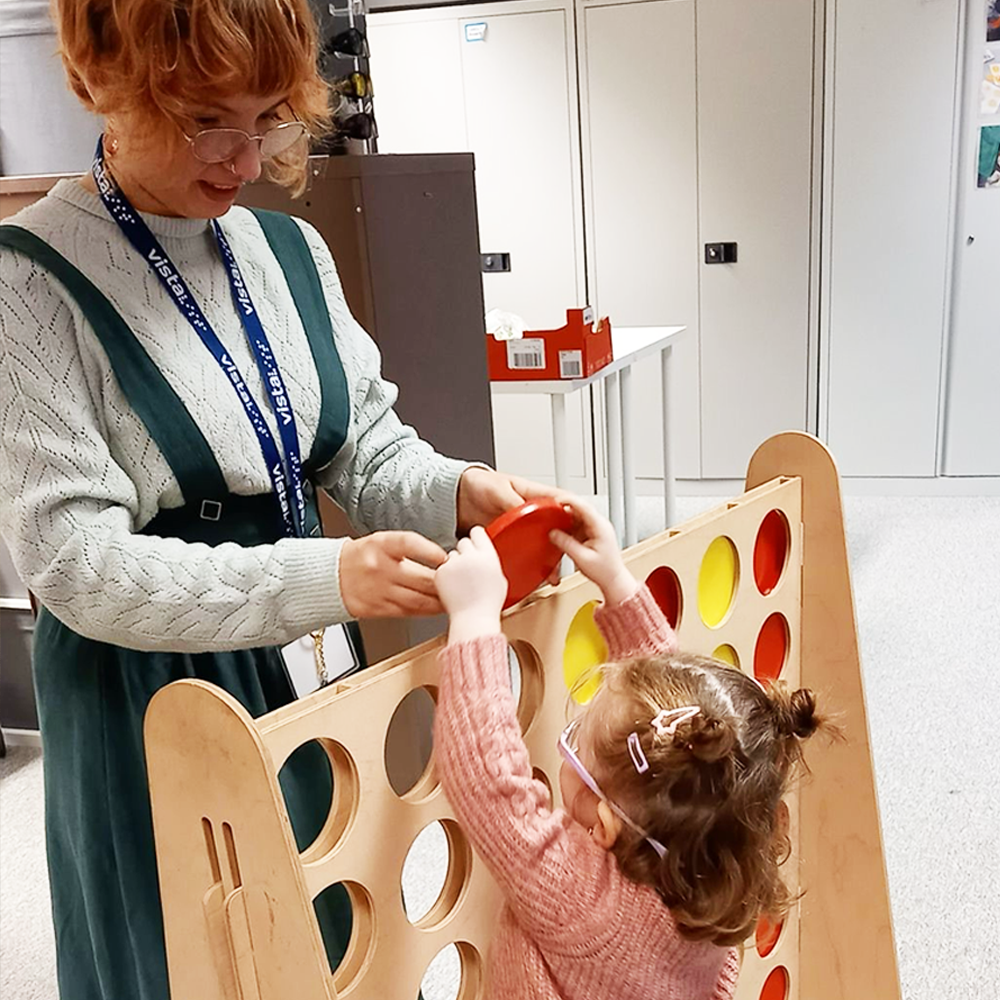 [Image Description: A woman wearing a Vista Staff Lanyard smiles at a young girl wearing glasses as they play large Connect Four together. Both of them are holding the game piece together as they place it in the slot at the top of the grid.]