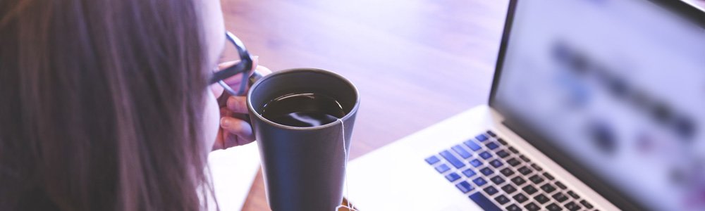 [Image Description: A woman sat holding a cup of tea whilst reading from a laptop screen. The contents of the screen are blurred so we cannot make them out.]