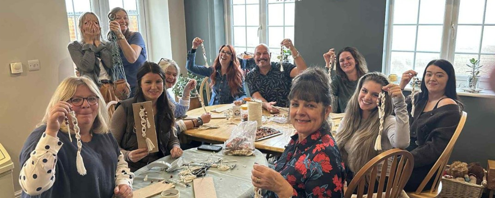 [Image Description: A group of people enjoying a macramé-making fundraiser. They smile and proudly hold up their finished keyrings.]