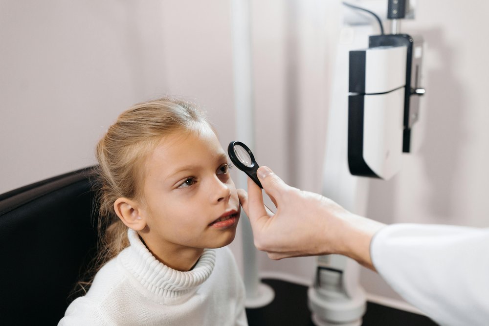 A young girl looks through a lens in an eye test room. Test equipment can be seen in the background.