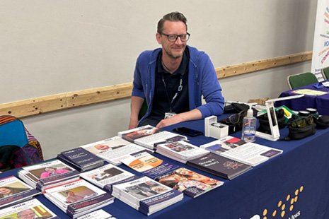 [Image Description: A man wearing glasses and a blue hoodie sits behind a Vista information table at an indoor event. The table is covered with brochures, leaflets, and assistive devices, and a navy cloth with the Vista logo and the tagline "Improving lives of people with sight loss." A banner for a nearby support service is partially visible in the background.]