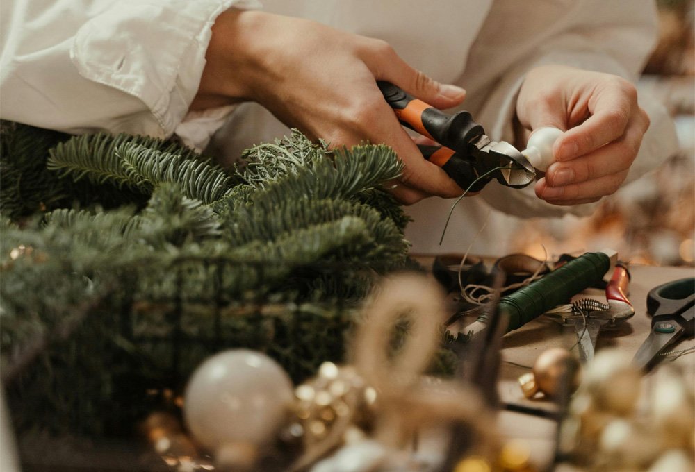 Image Description: A person uses a pair of cutters to cut a ribbon on a bauble. Pieces of foliage and other festive decorations and tools are on the table the person works at.