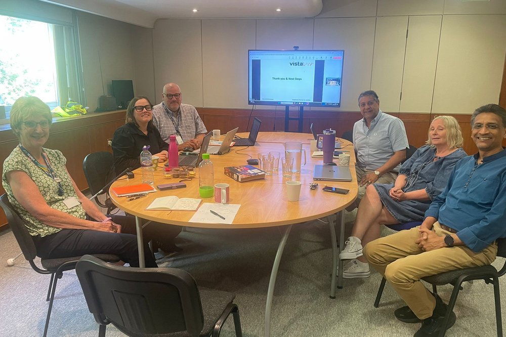 Image Description: Six people sit around a large meeting table in a well-lit room, smiling at the camera. Laptops, notebooks, drinks, and snacks are spread across the table. A screen in the background shows a Vista presentation slide titled “Thank you & Next Steps.”