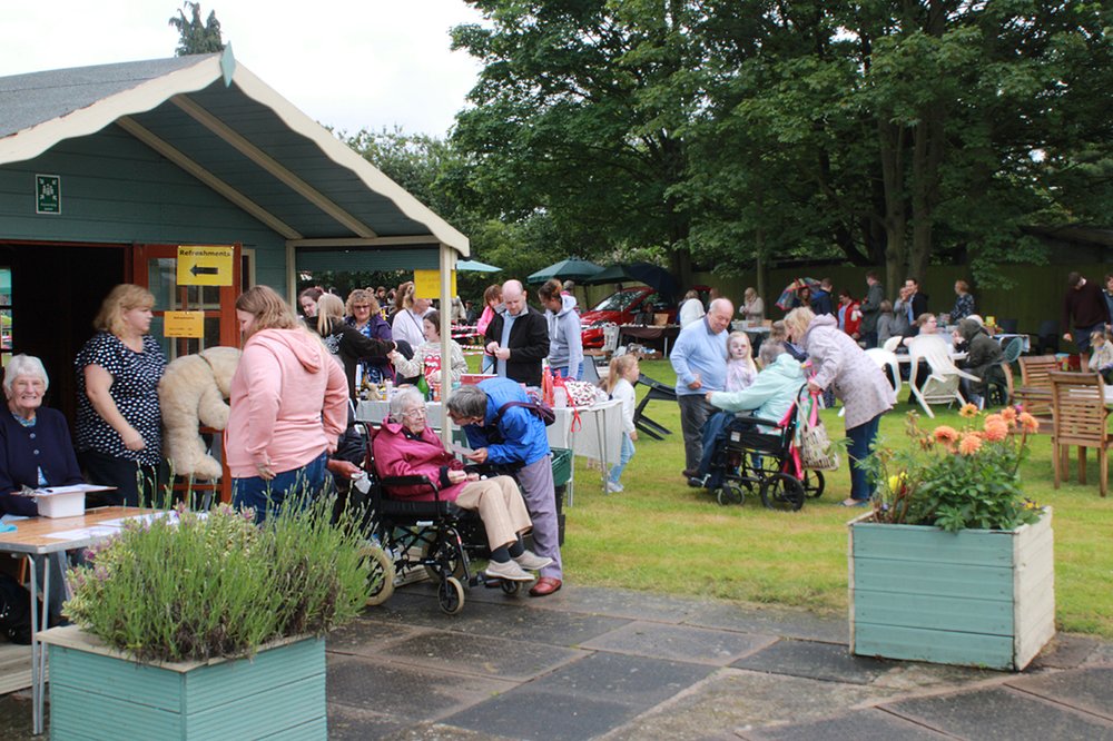 The kathleen Rutland garden, with large, green shed, with people milling about, some at a table playing games, other stood talking with ice creams