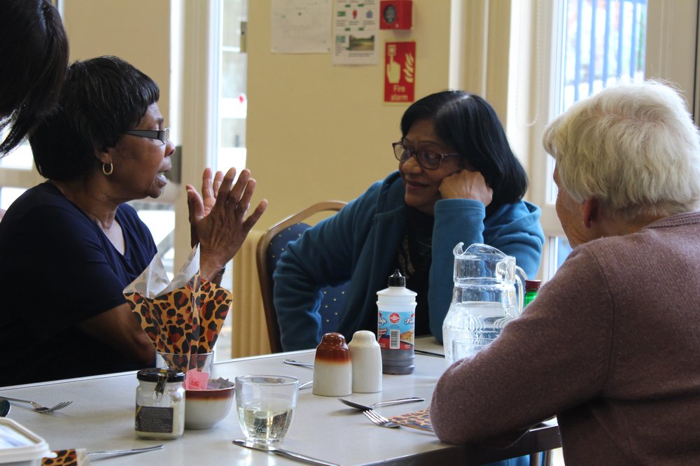 A picture of a lady helping another lady up from her chair.