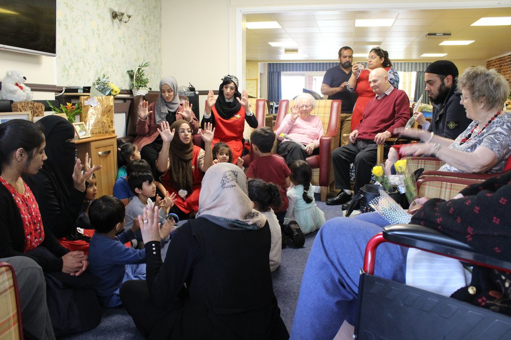 A group of children sat on the floor with their teachers with older residents in arm chairs around, laughing along.
