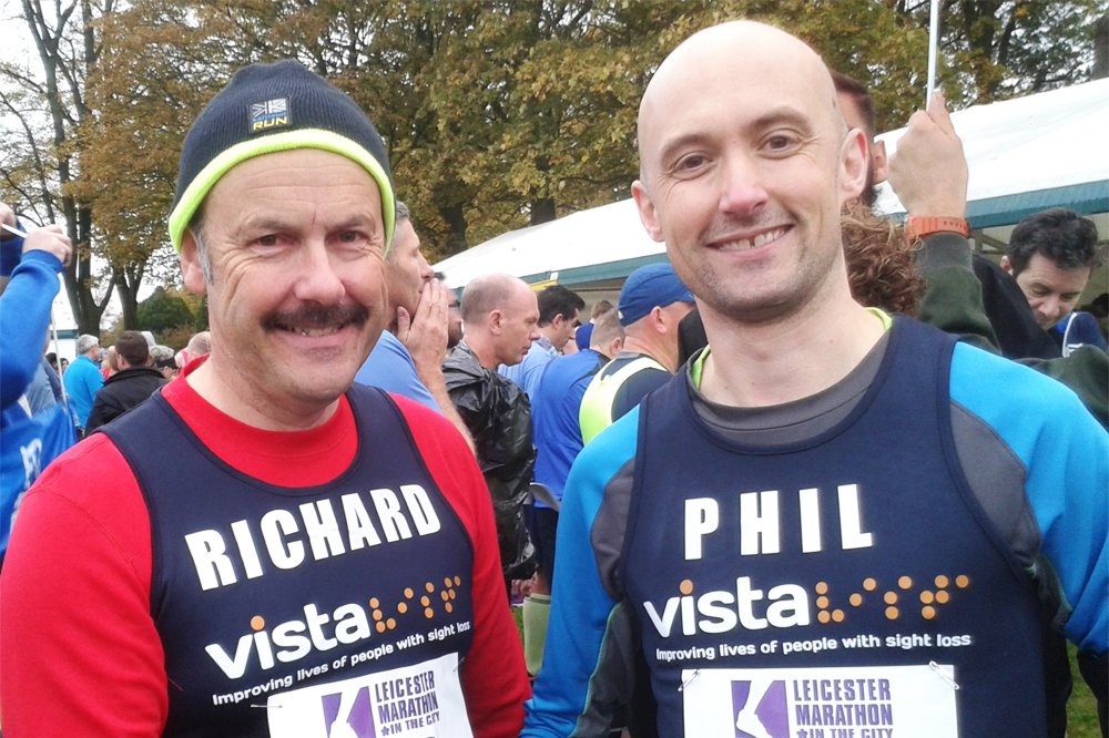Two men wearing Vista running bibs adorned with the names 'Richard' and 'Phil' stand smiling at a running event.