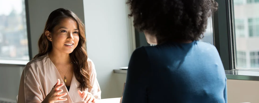 [Image Description: A woman sits across from another woman at a table, smiling at her as they talk.]