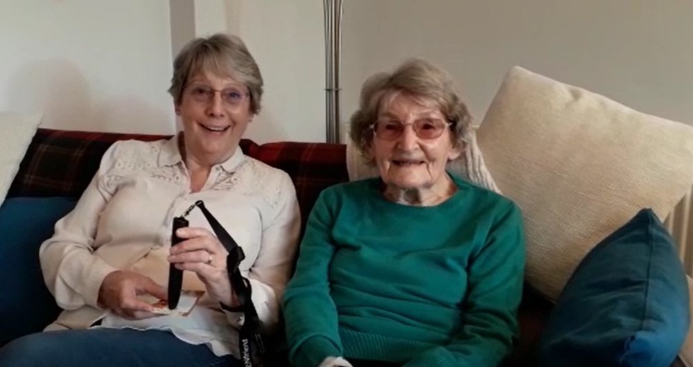 Image is of Jo and Mary are smiling and sitting on a sofa. Jo is holding an RNIB PenFriend.