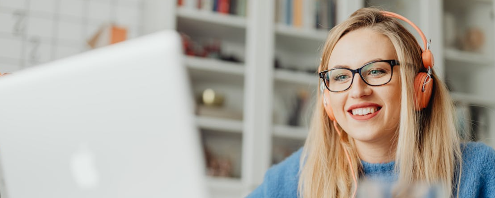 [Image Description: A woman wearing glasses and a pair of headphones smiles whilst looking at a laptop]