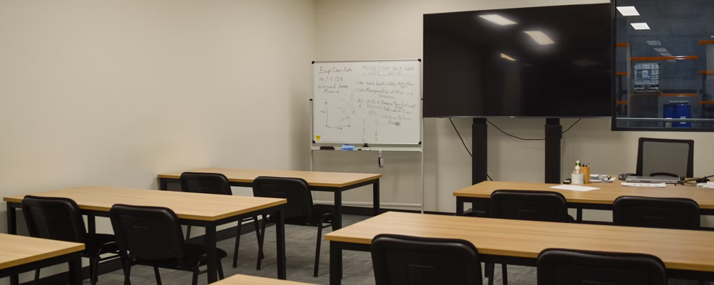 [Image Description: An empty classroom with rows of empty tables and chairs visible, and a television screen]