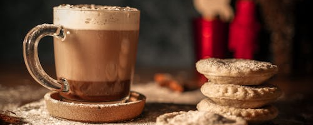 [Image Description: Three mince pies are stacked alongside a singular mince pie opposite a mug which has hot chocolate with a layer of cream on it. They are sat on a board which has been dusted with icing sugar.]