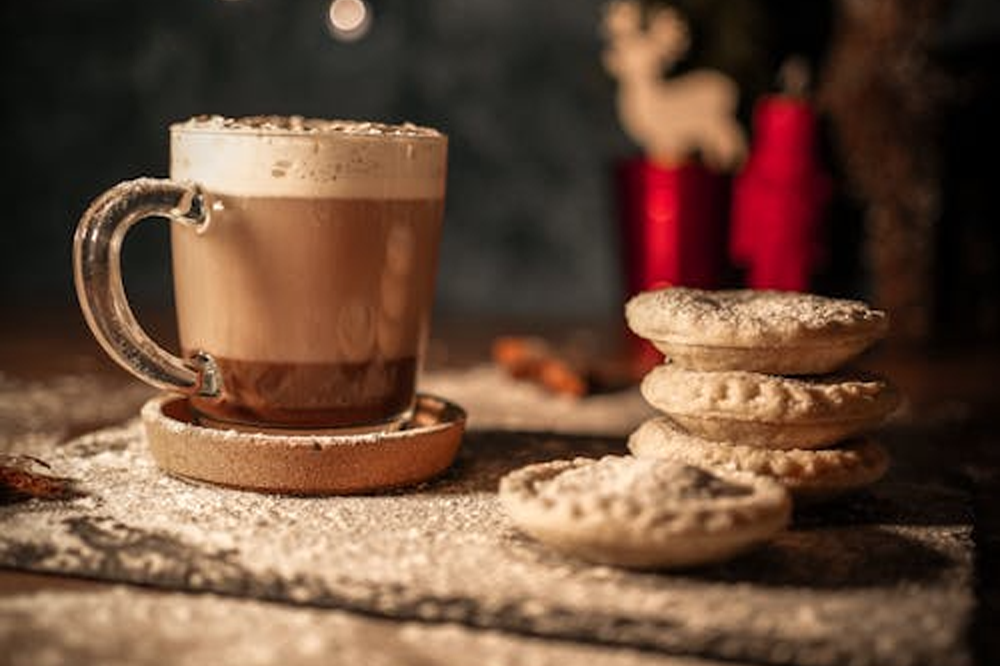 [Image Description: Three mince pies are stacked alongside a singular mince pie opposite a mug which has hot chocolate with a layer of cream on it. They are sat on a board which has been dusted with icing sugar.]