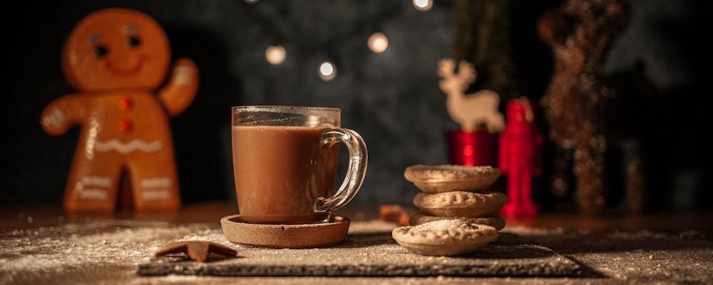 [Alt Image: A glass of hot chocolate with a stack of mince pies, set in front of festive holiday decorations and twinkling lights.]