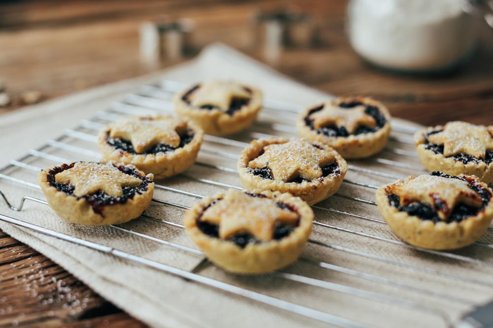 [Several mince pies sit on an oven rack with paper towel underneath them. The Mince Pies have stars for the top of their casings. A wooden worktop can be seen behind them.]