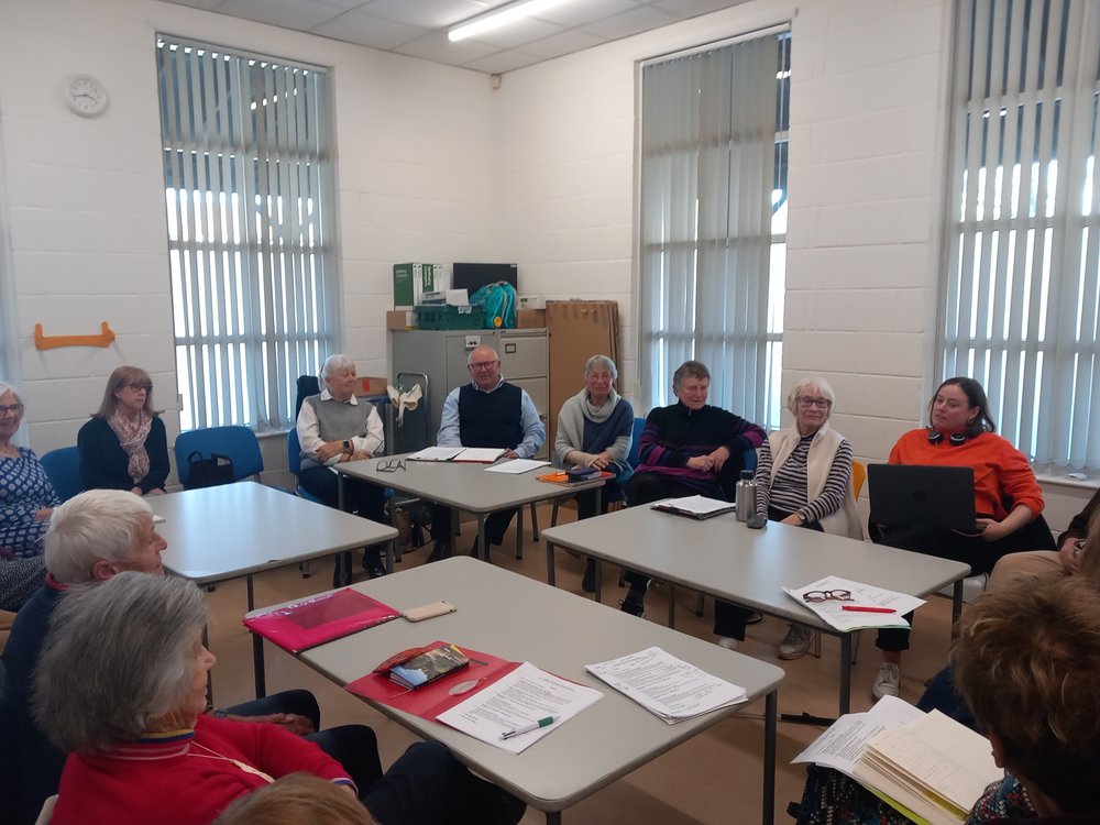 [Image description: The volunteer team are seated around central tables in a community room.  They are all looking and smiling at the camera.]