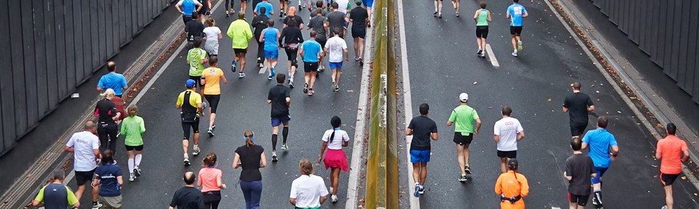Image Description: A group of people wearing various brightly coloured clothes run along a road together in an organised Marathon event.