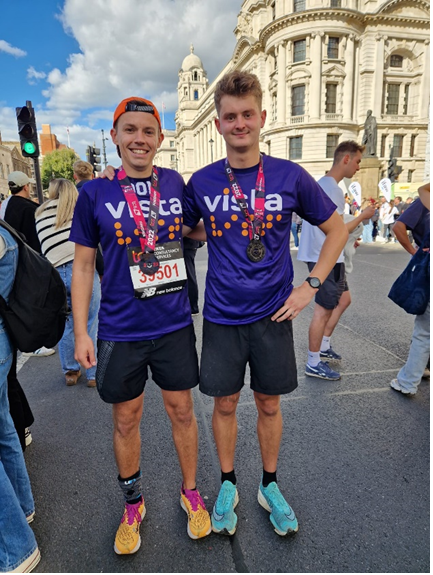 Image description: Two men standing smiling wearing Vista t-shirts with medals around their necks after completing a race. They stand in a street, with buildings and people behind them.