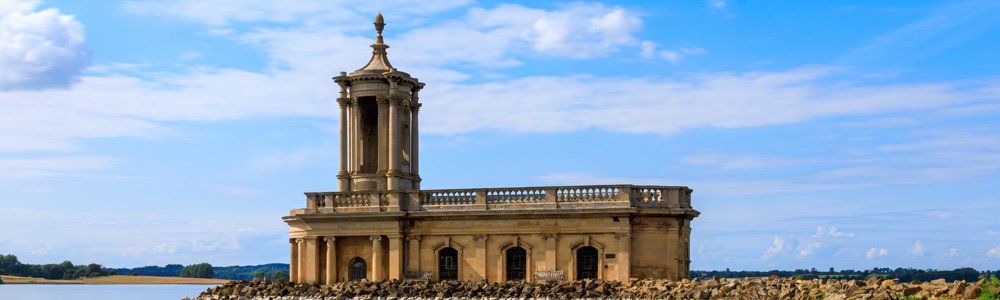 Image Description: Normanton Church at Rutland Water. The church is sat on the shoreline, surrounded by rocks. Water can be seen against the shore, and in the background more of Rutland Water Park can be seen.