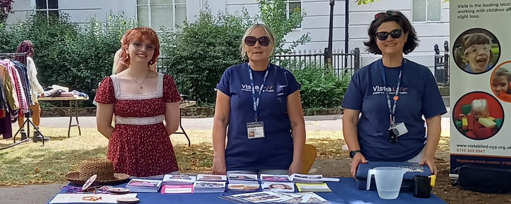 [Image Description:  Three people stand smiling behind a Vista-branded table at an outdoor community event. The table is laid with leaflets and resources, with a “COME MAKE FRIENDS WITH VISTA” sign on the ground. A Vista pop-up banner stands to the side under a tree providing shade.]