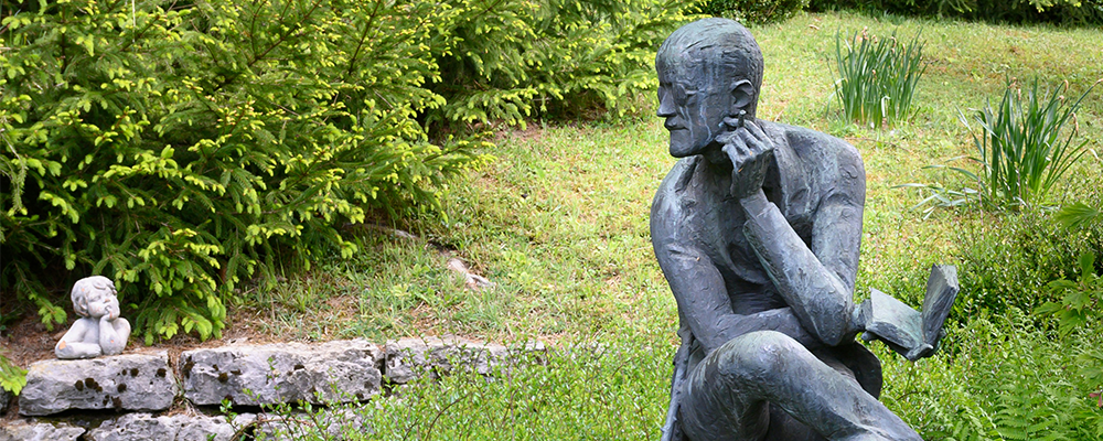 [Image Description: A contemplative metal statue sits in a lush garden, legs crossed and chin resting on its hand as if deep in thought.]