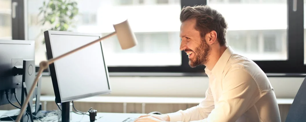[Image Description: Man in White Dress Shirt Sitting on Black Rolling Chair While Facing Black Computer Set and Smiling]
