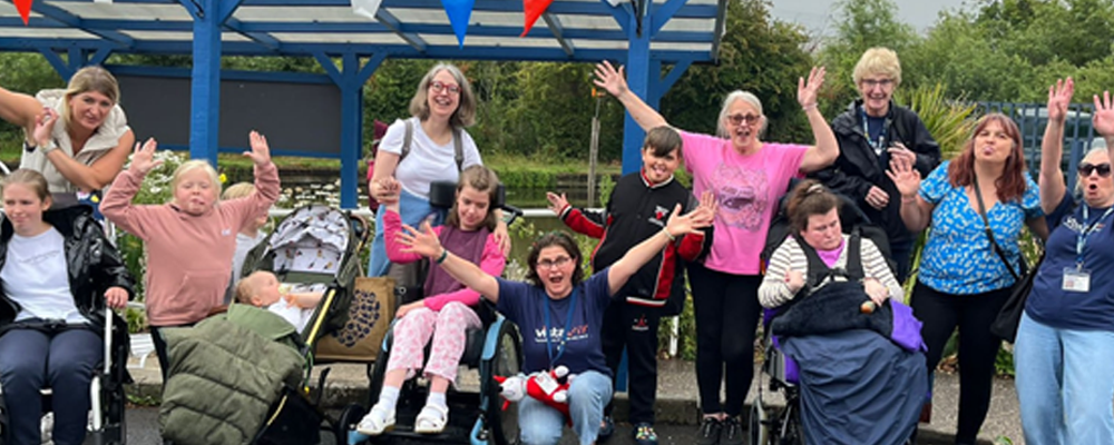 [Image Description: Families and staff pose by the canal with arms raised and big smiles. Some are in wheelchairs or holding little ones. Grey skies but high spirits.]