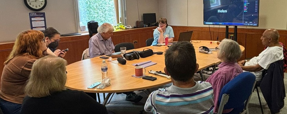 [Image Description: This image shows a group of eight people sitting around a large oval table in a meeting room. They’re talking and looking at phones or papers. In front of them is a TV screen displaying a Zoom call, where more people have joined remotely. The table has mugs, water bottles, and some tech devices on it. The atmosphere looks friendly and focused.]