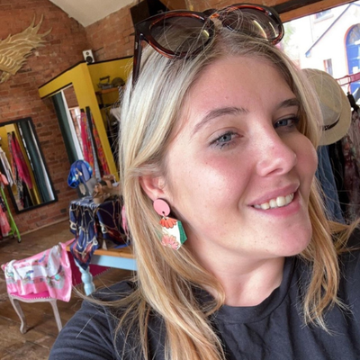 Tilly is smiling at the camera, with her clothes and accessories shop in the background. Shas blonde hair and dark sunglasses resting on top of her head.