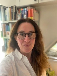 A photo of a woman from shoulder height. She's looking at the camera, wearing dark framed glasses and a white shirt. She has long brown hair. Behind her are some bookshelves to the ceiling and a the room has white walls.