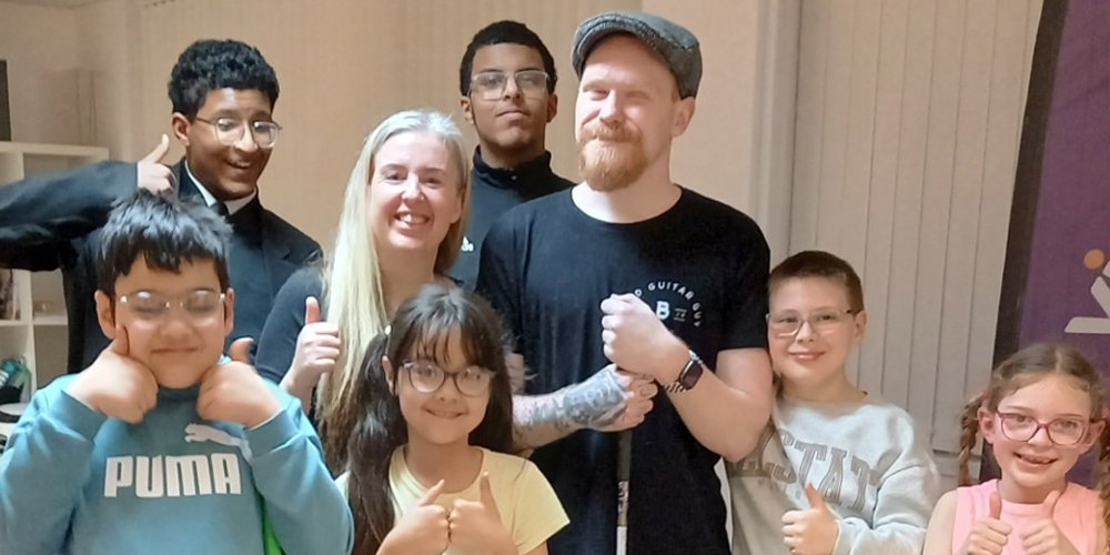 Vista Ambassador Craig, the Blind Guitar Guy, is surrounded by young people who are giving thumbs up towards the camera and smiling.