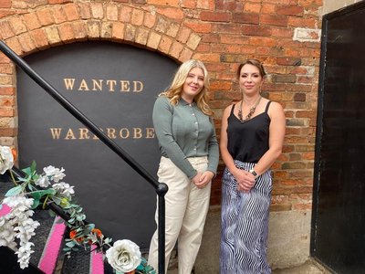Tilly , left and her business partner and friend Victoria, right, stand at the bottom of the stairs outside their shop, Wanted Wardrobe. Flowers decorate the bannisters and Wanted Wardrobe is on the black painted wall behind them.