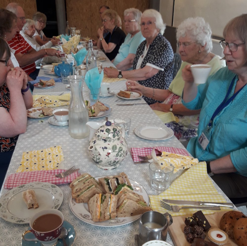 a table full of cakes and sandwiches, with people either side tucking in