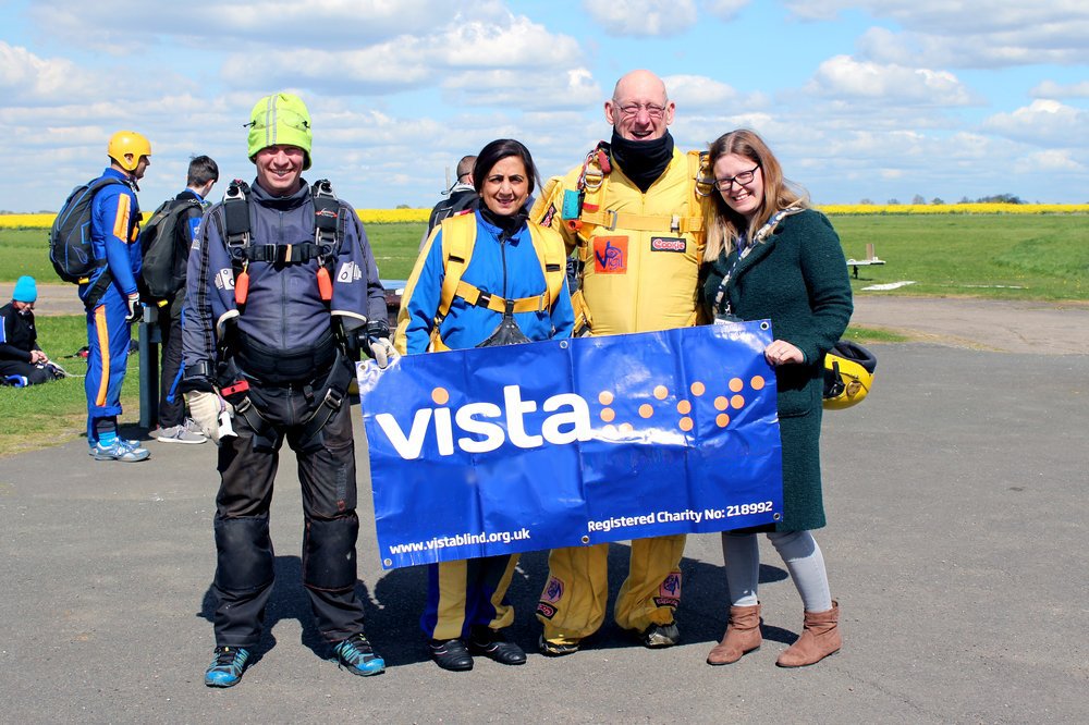 Image description: two men and two women stand on the airfield holding a large Vista banner. They are completing a skydive.